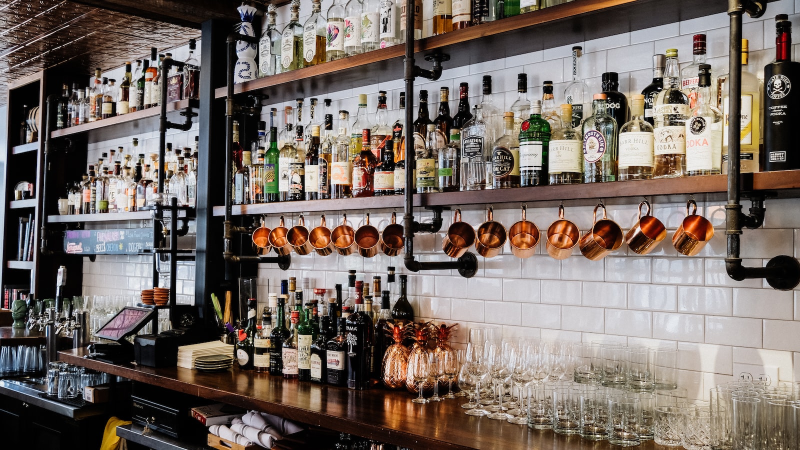 A well-stocked bar is pictured, with shelves and cabinets filled with bottles of various liquors, mixers, and tools. The bar is adorned with an array of glassware, including cocktail glasses, wine glasses, shot glasses, and beer glasses. The glasses are arranged neatly on shelves and hanging racks, ready for use. The lighting in the room casts a warm glow, adding to the cozy and inviting ambiance of the bar.