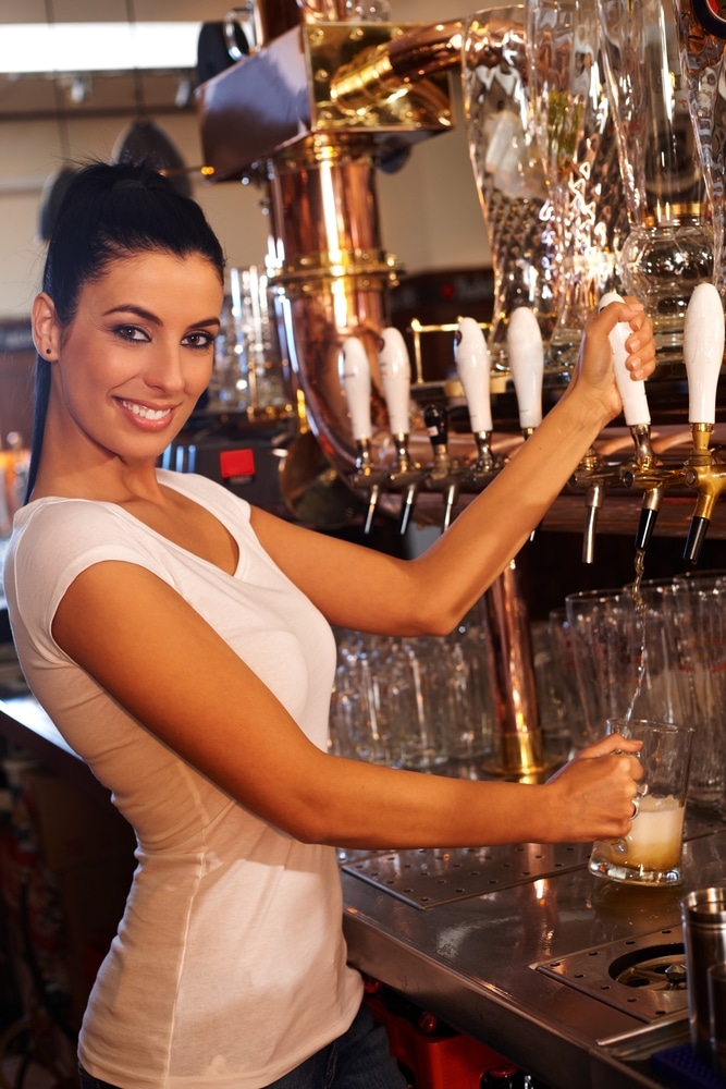 A female bartender stands behind a bar pouring a beer.